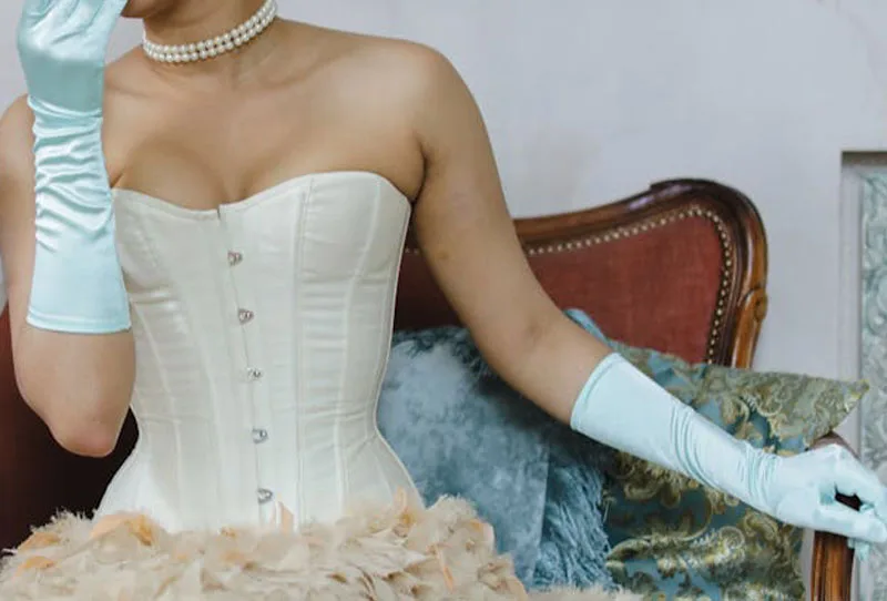 Femme en corset blanc long, jupe crème à froufrous et gants bleu ciel, assise dans une pose élégante.